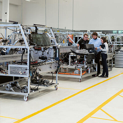 Drei Ingenieur:innen stehen in einer Produktionshalle an einem Tisch mit Geräten und einem Laptop.