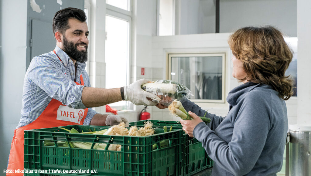 Ein Mann mit Schürze und Hygiene-Handschuhen gibt einer Frau ein Paket Gemüse über die Theke.