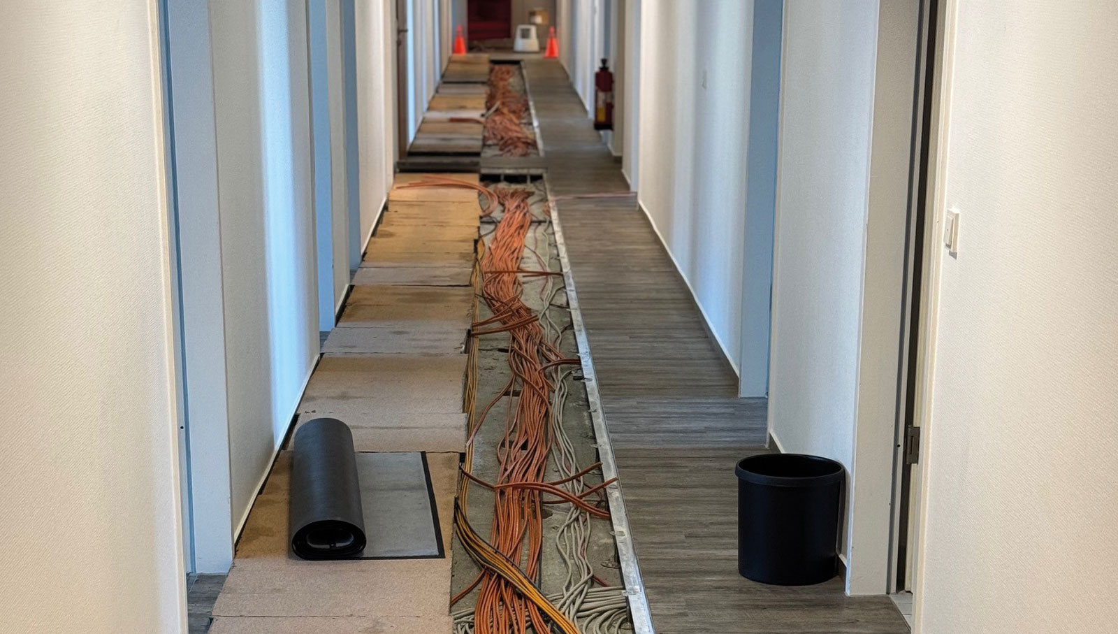 Langer Flur bei dem die Bodenplatten in der Mitte den kompletten Gang runter aufgerissen sind und viele lange orangene Kabel freigelegt zeigen. An der Seite noch ein aufgerollter Teppich und ein Mülleimer.