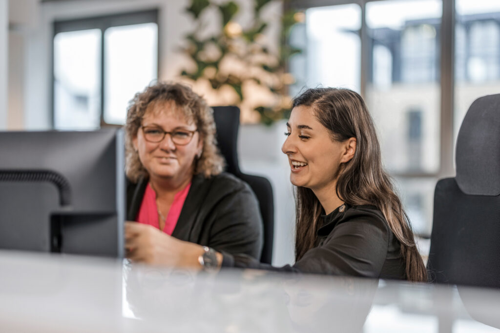 Eine Mitarbeiterin erklärt einer anderen Mitarbeiterin etwas sitzend am Bildschirm.