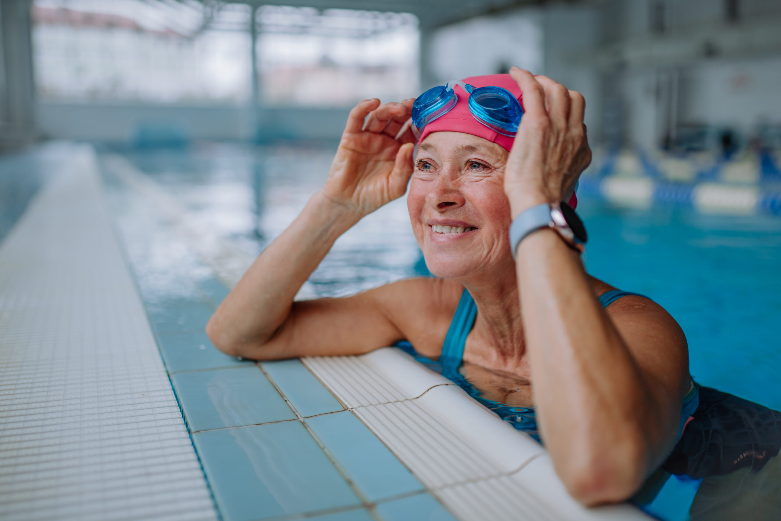 Ein ältere Frau im Pool mit Badekappe lehnt lächelnd an den Pool-Rand, hat sich gerade die Schwimmbrille auf die Stirn geschoben.