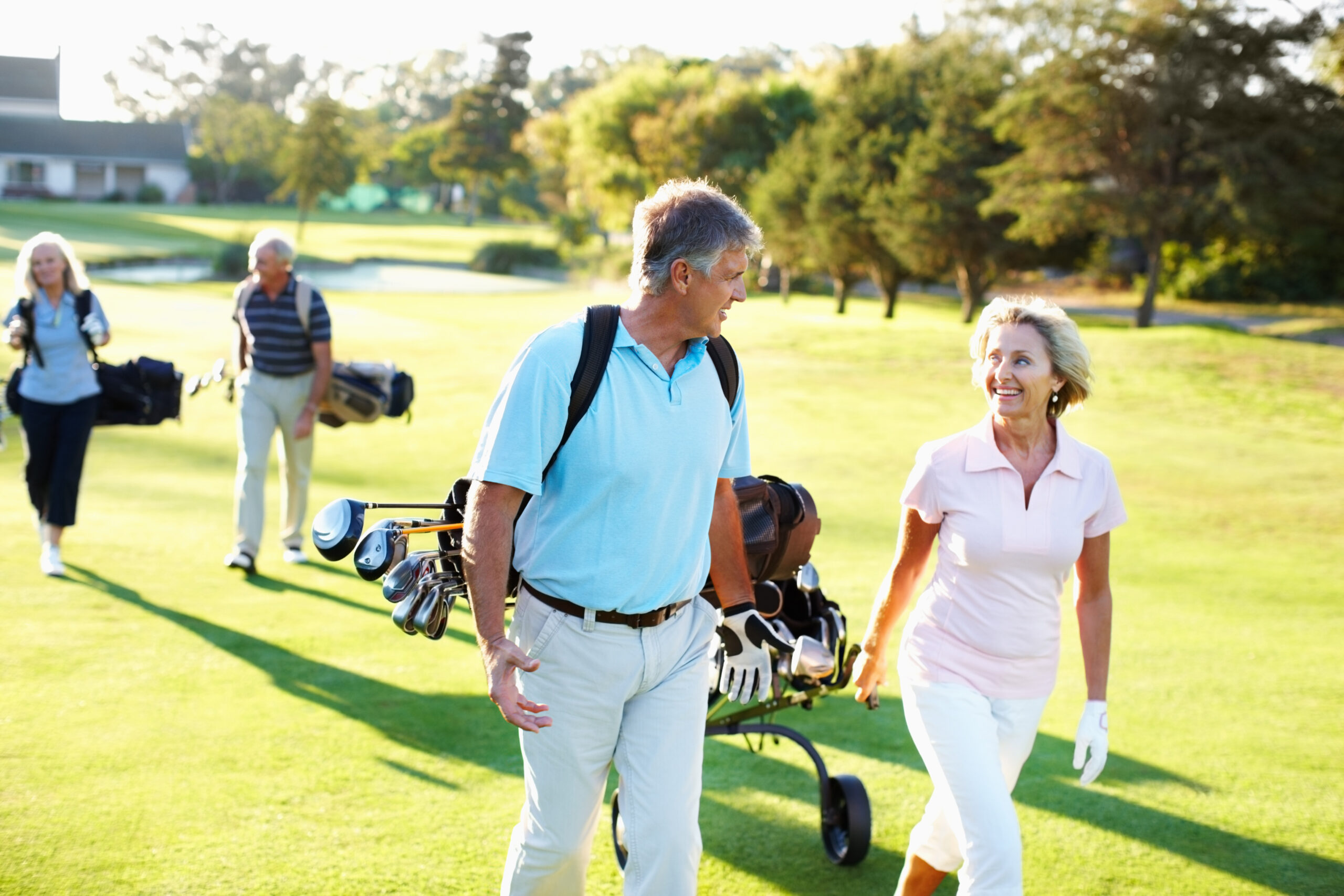 Ein älteres Pärchen geht lächelnd über den Golfplatz. Er trägt eine Golftasche und zieht einen Caddy hinter sich her. Hinter ihnen ein weiteres Pärchen.