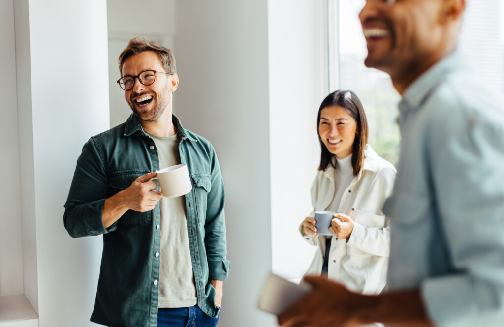 3 Menschen in Business Casual lachen stehend zusammen während einer Pause mit Kaffee in der Hand.