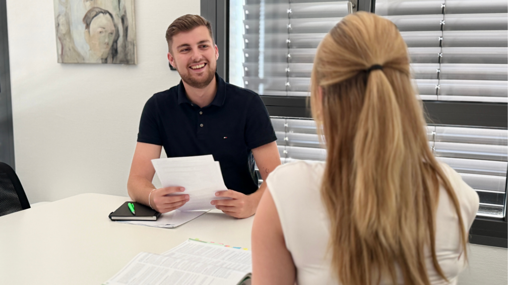 Gespräch in einem Büro an einem Besprechungstisch. Eine Person präsentiert Dokumente einer gegenüber sitzenden Person. Auf dem Tisch liegen Arbeitsunterlagen, ein Notizbuch und ein Stift; im Hintergrund befinden sich große Fenster mit Jalousien.