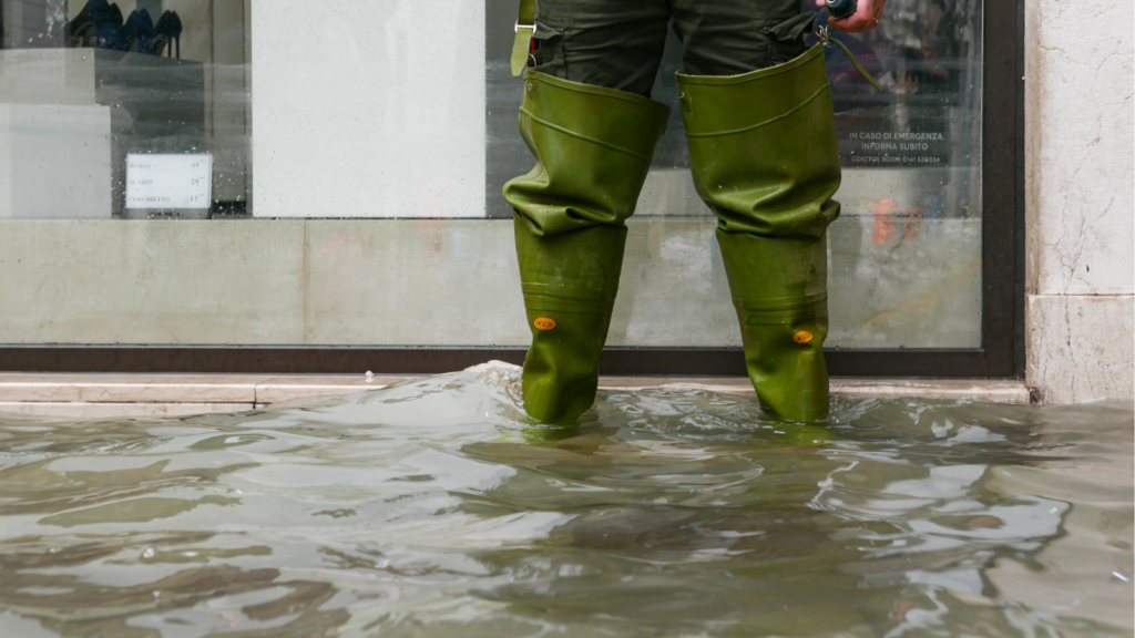 Person in grünen Gummistiefeln steht knietief in überflutetem Wasser vor einem Gebäude. Das Bild veranschaulicht die Folgen von Hochwasser und Starkregen für Immobilien.