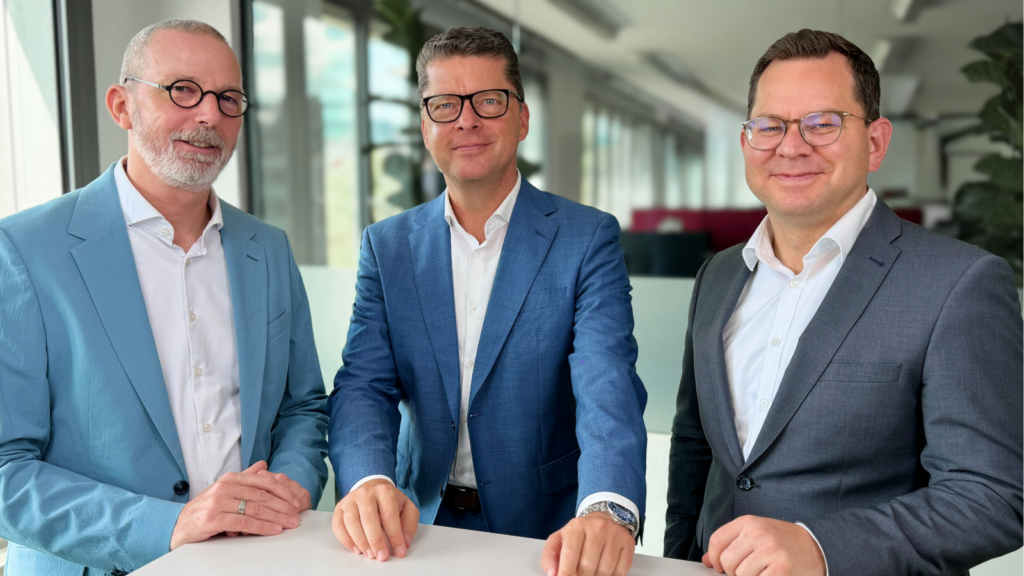 Gruppenporträt von drei Personen an einem weißen Stehtisch in einem modernen Büro. Die Personen tragen Hemd und Sakko und stehen nebeneinander vor einer unscharfen Büro-Kulisse mit Fenstern und Pflanzen. Die Szene vermittelt ein professionelles Arbeitsumfeld.